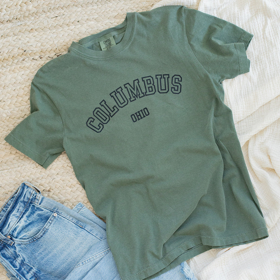 Green t-shirt with 'Columbus' text and blue jeans on a textured surface