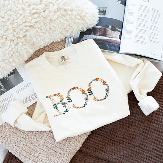 Embroidered 'BOO' design on a white fabric with books and a fluffy white pillow in the background.