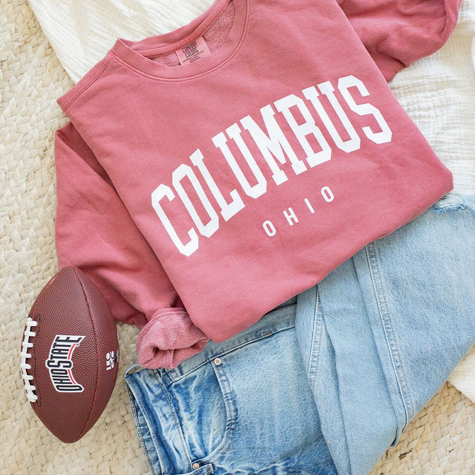 Pink sweatshirt with 'Columbus, Ohio' text, blue jeans, and football on a beige surface.