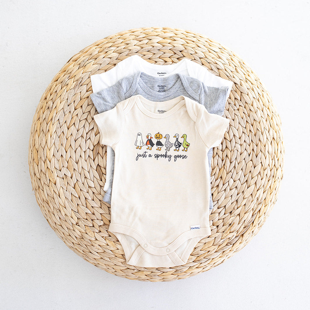 Three baby bodysuits in white, light gray, and cream are stacked on a round woven straw mat against a white background. The front bodysuit is cream with a graphic of cartoon geese and the text just a spooky goose"