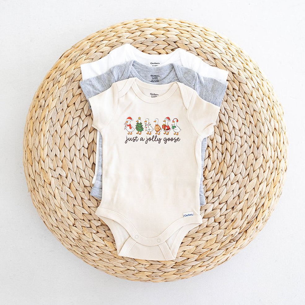 A cream-colored baby bodysuit with a Christmas graphic and the text 'just a jolly goose' is laid flat on a round woven straw mat, with two gray baby shirts partially visible behind it against a white background.