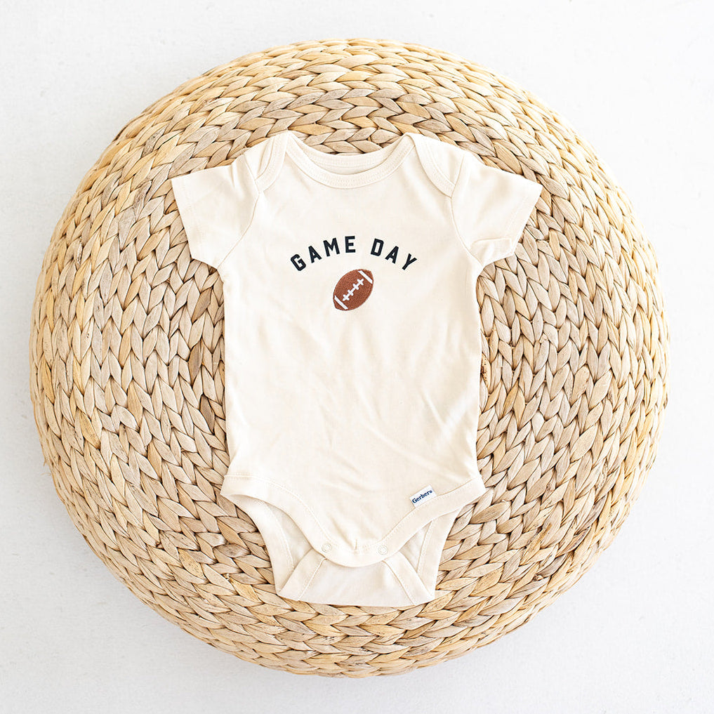 A cream-colored baby bodysuit with printed 'GAME DAY' and a football graphic embroidered on the chest, laid flat on a round woven straw mat against a white background.