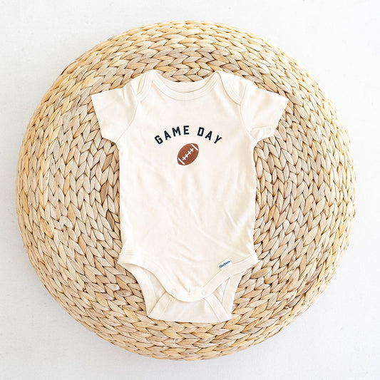 A cream-colored baby bodysuit with printed 'GAME DAY' and a football graphic embroidered on the chest, laid flat on a round woven straw mat against a white background.