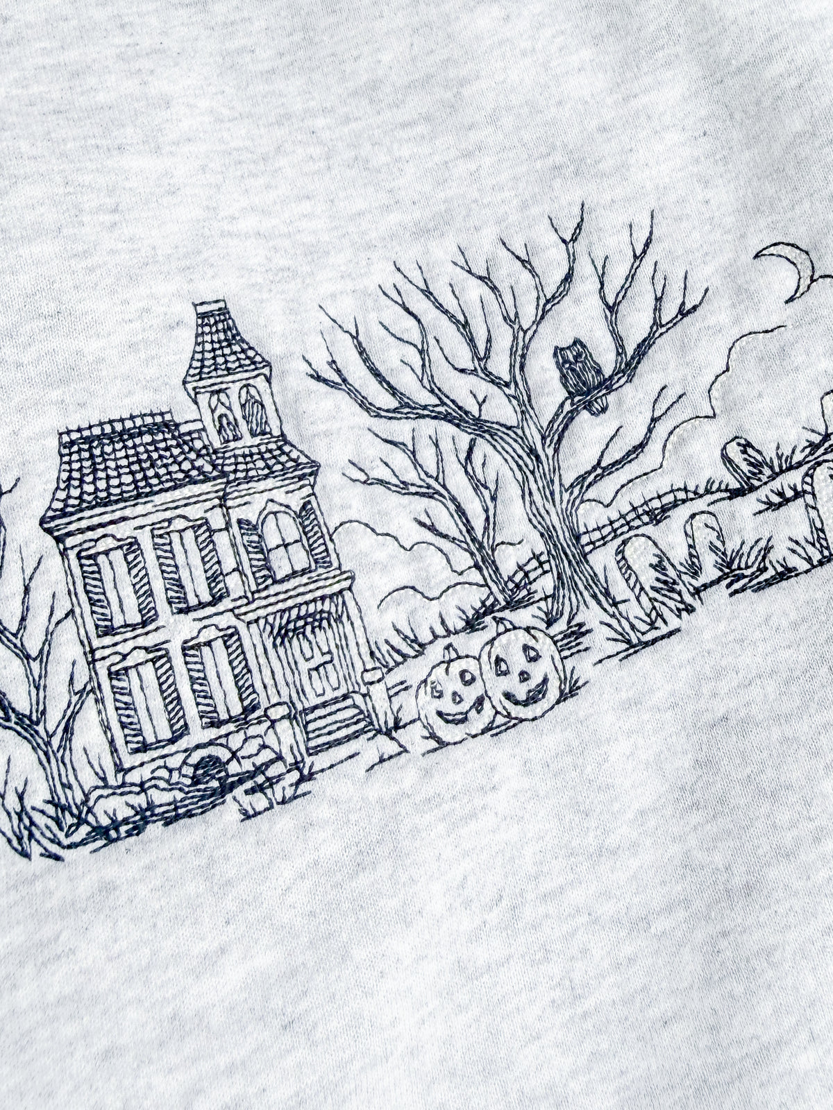 Close-up of a light gray heather fabric embroidered with a black line art illustration of a haunted house, bare tree, jack-o'-lanterns, and an owl.