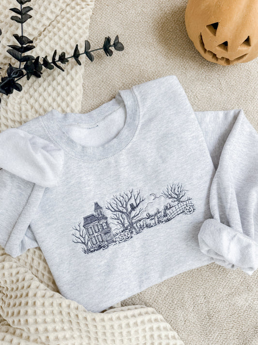 A light heather gray long-sleeve sweatshirt with embroidery of a haunted house, bare trees, and a pumpkin patch in a flat lay on a cream knit blanket with a jack-o'-lantern and eucalyptus sprig.