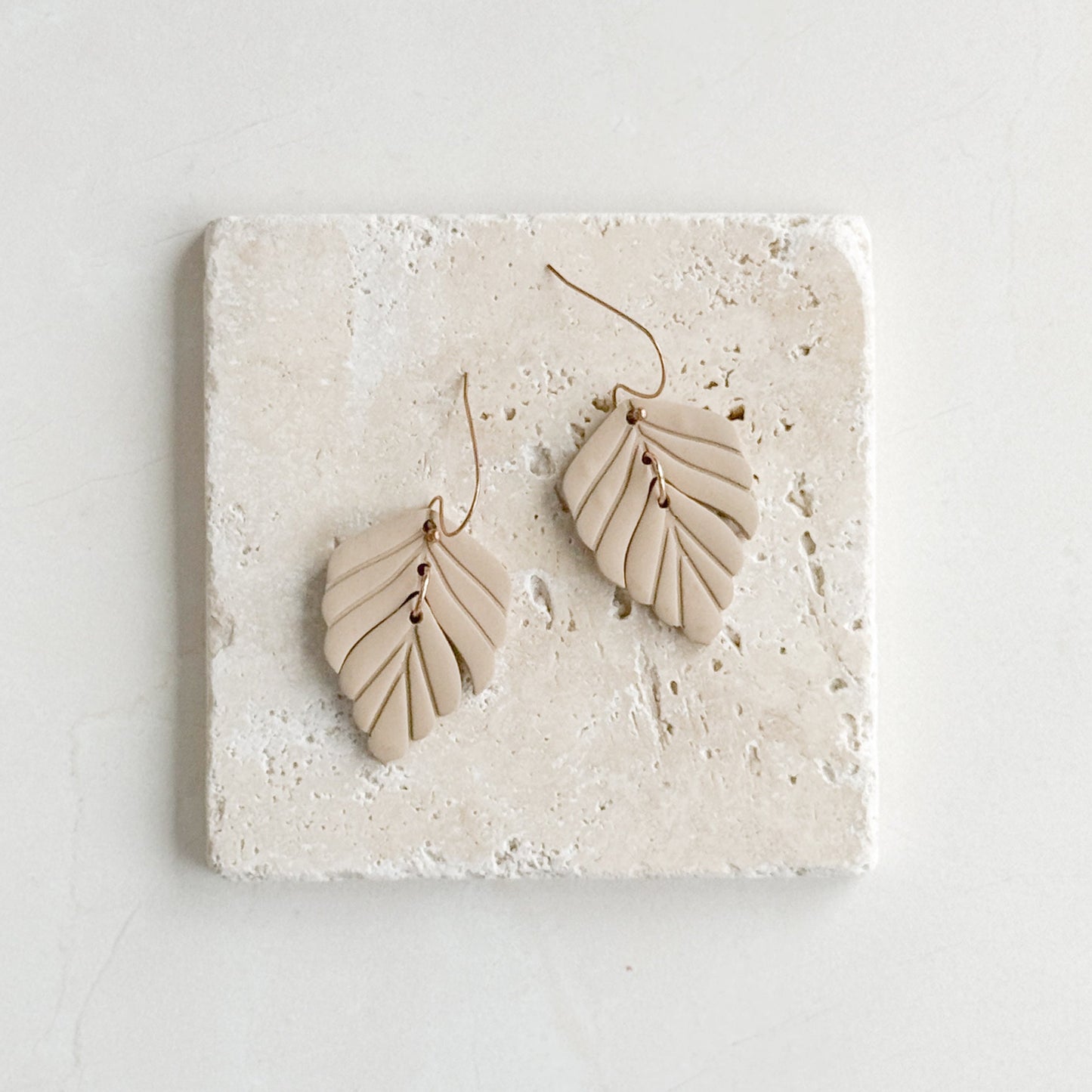 Pair of beige feather-shaped earrings on a textured stone surface with a light gray background