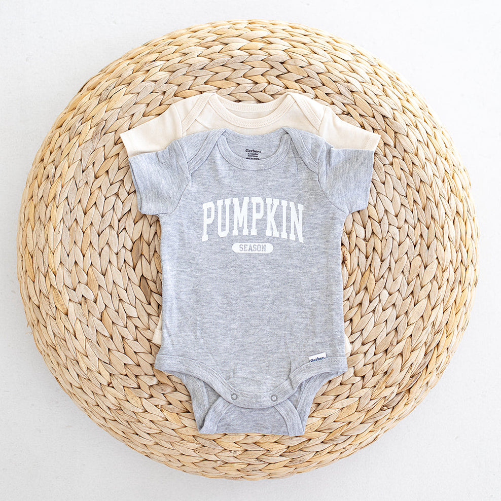 Two baby bodysuits, one heather gray with pumpkin season printed in white and one cream, arranged on a round woven straw mat against a white background.